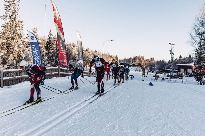 The image shows a group of people cross-country skiing on a snowy trail, with participants wearing various winter sports gear, surrounded by snow-covered trees and a winter sports event banner.AI-genereret indhold kan være ukorrekt.