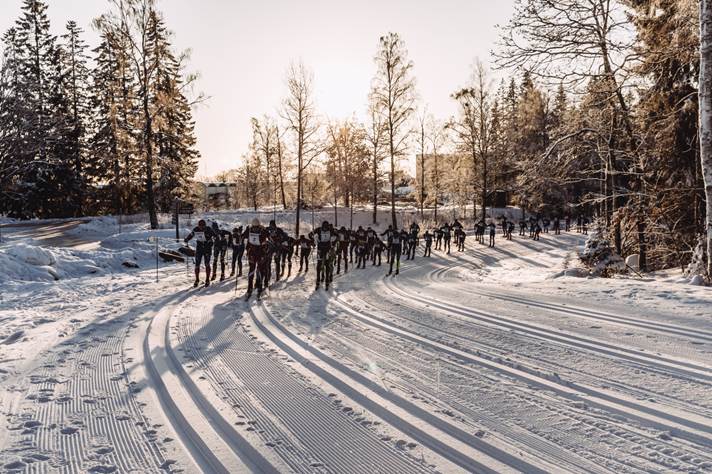 The image depicts a group of people cross-country skiing on a snowy trail, surrounded by a forest of tall, snow-laden trees.AI-genereret indhold kan være ukorrekt.